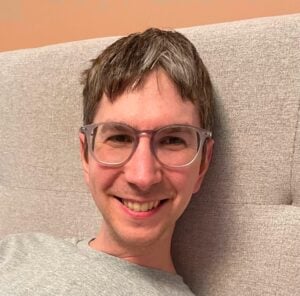 Portrait of Derek, white face with broad smile and glasses.